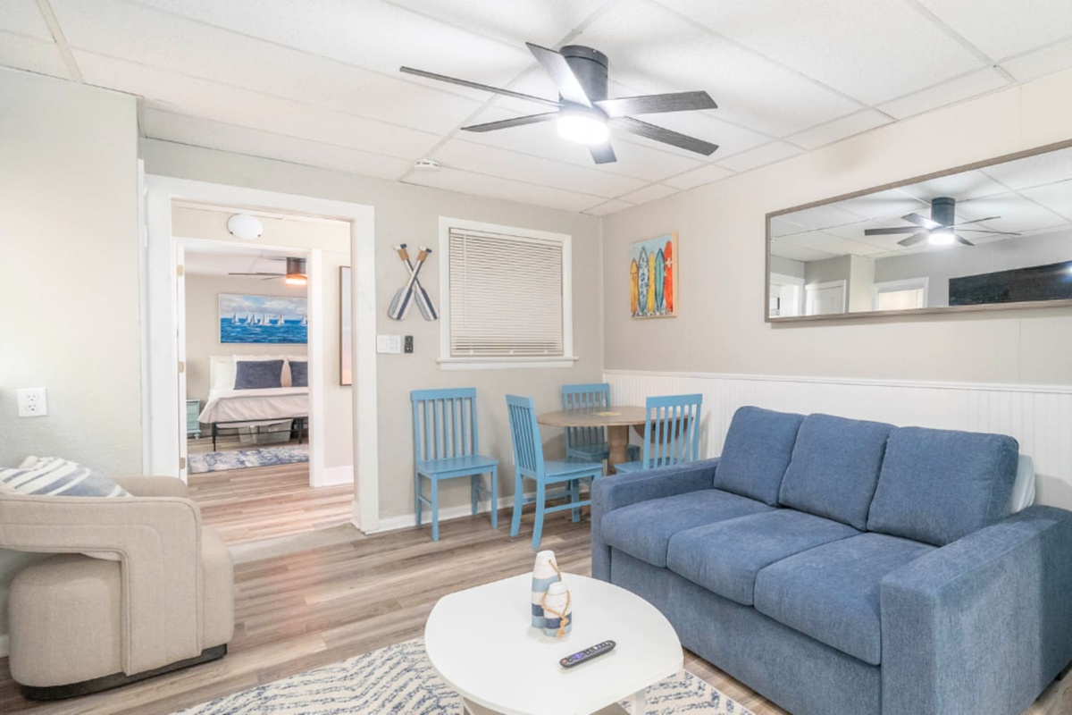 Bright coastal living area with a comfortable sofa, armchair, and dining nook for easy meals. Ceiling fan and soft lighting keep the space cool and relaxed, with an open flow to the bedroom for a comfortable stay.