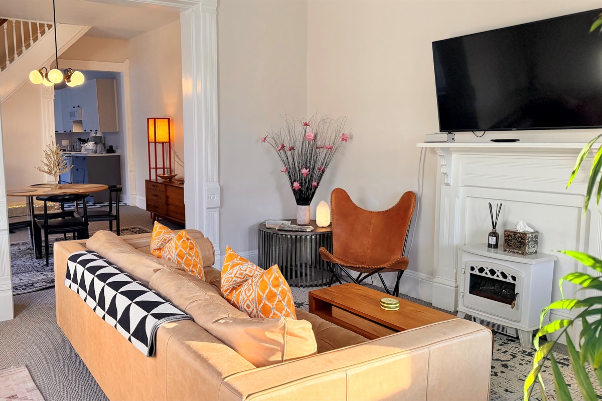 Living room with a leather sofa, two chairs, area rugs, lamps, and electric fireplace beneath a mounted TV. The front door opens into this room, which flows through the dining area, kitchen, laundry room and out the back door to the yard and parking.