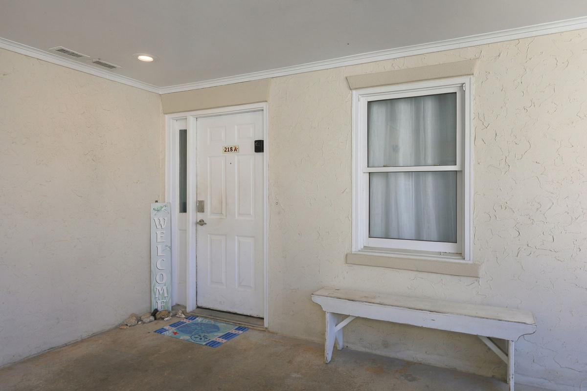 Covered entry area featuring a white front door, weathered wood bench, single-hung window with interior privacy shade, and a vertical coastal welcome sign.   