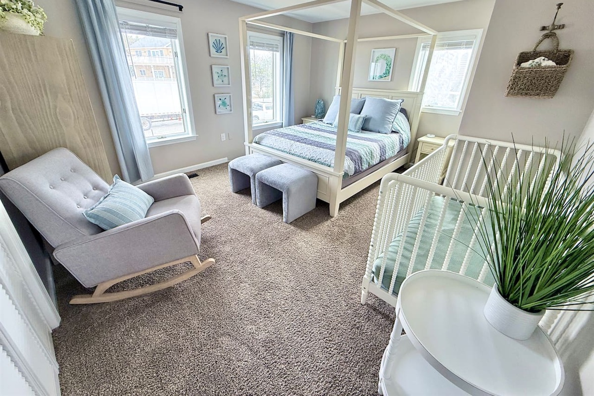 This family-friendly bedroom offers a relaxing, coastal retreat featuring a four-poster canopy bed, a white crib, and a cozy rocking chair. Light blue curtains and nautical art complete the space.