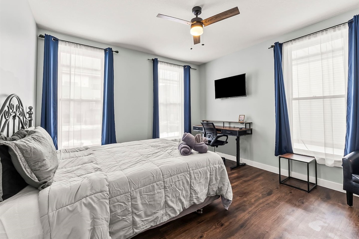 Bright bedroom view showcasing large windows, blackout curtains, ceiling fan, and a dedicated work desk.