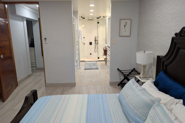 Primary bedroom at Villa Coral featuring a dark wood king bed frame, coastal blue bedding, and a direct view into the walk-in closet and glass steam shower in Dorado, Puerto Rico.

