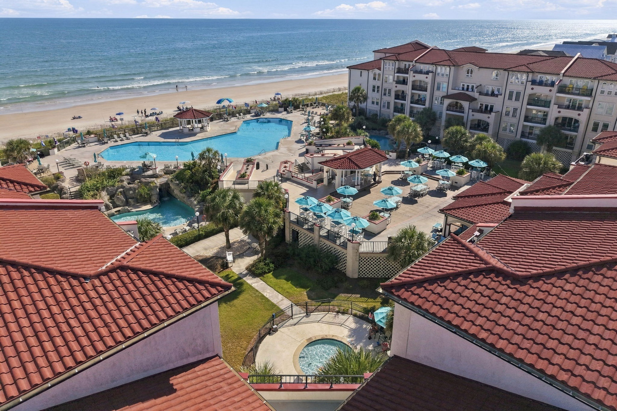 Elevated perspective framed by terracotta rooflines, overlooking the central pool courtyard, sun decks, landscaped walkways, and oceanfront shoreline.