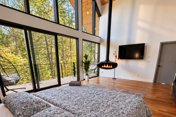 Living room: wood-burning fireplace glowing beside tall forest-view windows and a plush area rug. Cozy evenings in the trees.