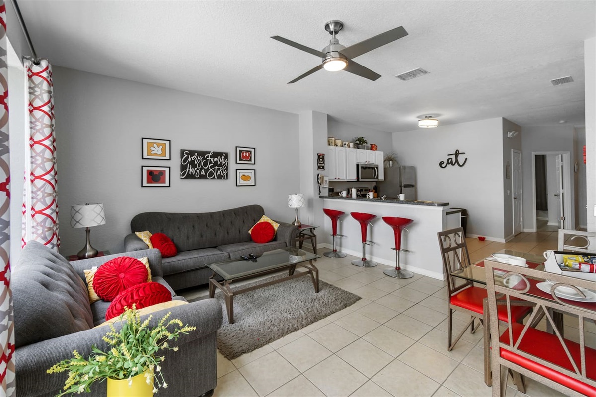 - Open living area with plush seating, bold red accents, and modern ceiling fan  
- Breakfast bar with red stools connects to a fully equipped kitchen nearby  
- Spacious layout ideal for relaxing, dining, and gathering with loved ones