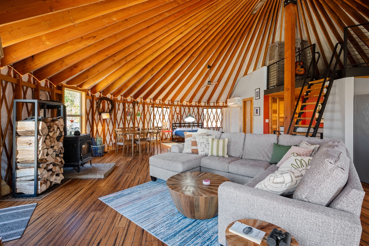 Open-concept living area with oversized sectional seating, wood-burning stove, vaulted ceilings, and panoramic yurt architecture.