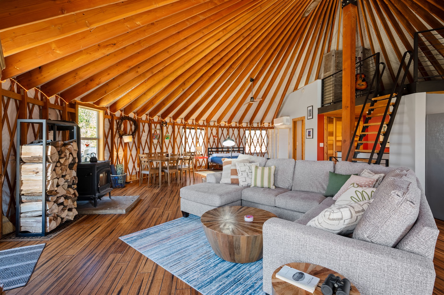 Open-concept living area with oversized sectional seating, wood-burning stove, vaulted ceilings, and panoramic yurt architecture.