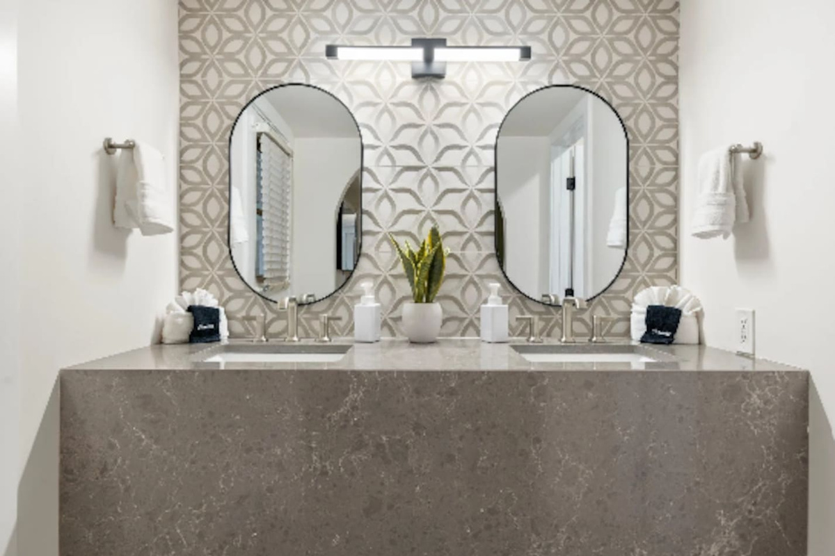 A dual vanity in patterned stone tile sets a spa-like tone, with a moody black-tiled shower just beyond. Every surface here feels intentional, right down to the oval mirrors and brushed fixtures.