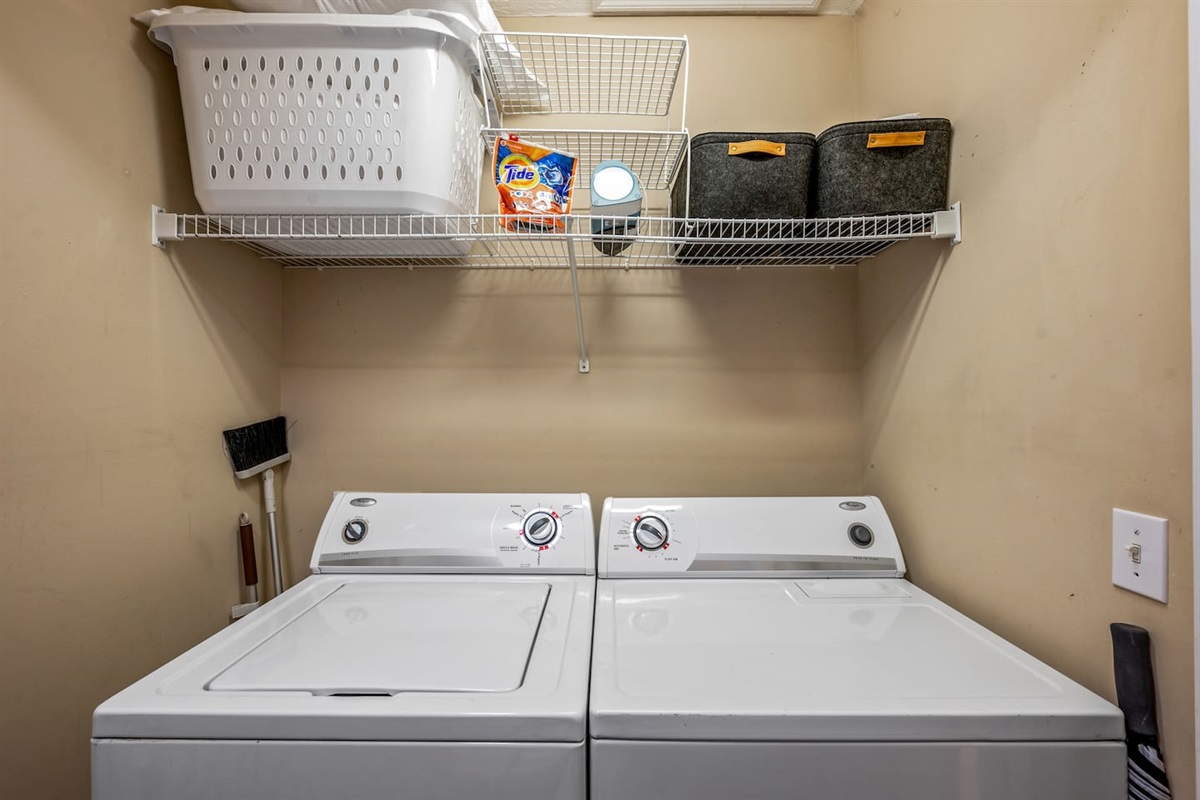 Convenient in-unit laundry area featuring a washer and dryer, with shelving above for storage. Essentials like detergent, laundry baskets, and cleaning supplies are provided for your stay.