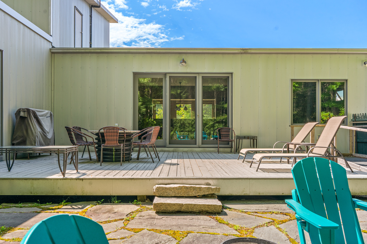 Modern design meets nature at "The Crag." Looking up from the fire pit patio, the full 33-foot deck features sun loungers, outdoor dining, and large Andersen doors—framed by striking corrugated siding and blue Maine skies near Acadia.