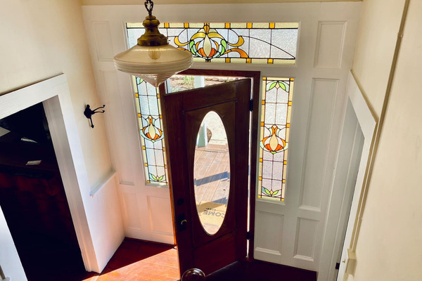 Afternoon light turns the stained-glass entry into a jewel box. The living room opens to the left; the first-floor primary suite—the "Boudoire de Bouge" and private bath—is tucked to the right. Historic charm, thoughtful flow.