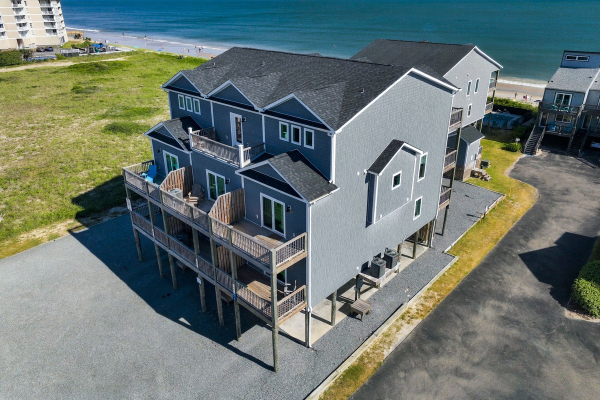 Elevated aerial view capturing the entire beachfront townhouse complex, highlighting the multi-tiered private balconies, dark shingle roofing, and footsteps-away access to the beach and ocean.   