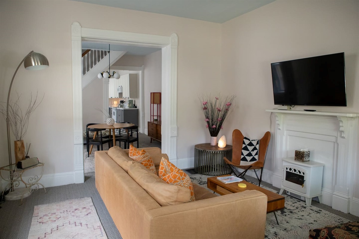 Living room with a leather sofa, two chairs, area rugs, lamps, and electric fireplace beneath a mounted TV. The front door opens into this room, which flows through the dining area, kitchen, laundry room and out the back door to the yard and parking.
