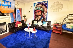 The living room is colorful with a striking mural, black sofas, a blue rug, and a fireplace. A hanging chair and red curtains add to the decor, while a small table with vases and a metal wall