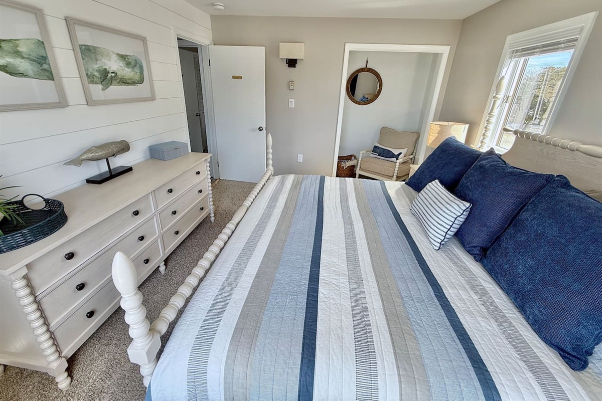 This coastal-themed bedroom features a white spindle bed with blue-striped bedding, a matching dresser with whale-themed decor, and a cozy reading nook, all coming together to create a relaxing, nautical-inspired escape.