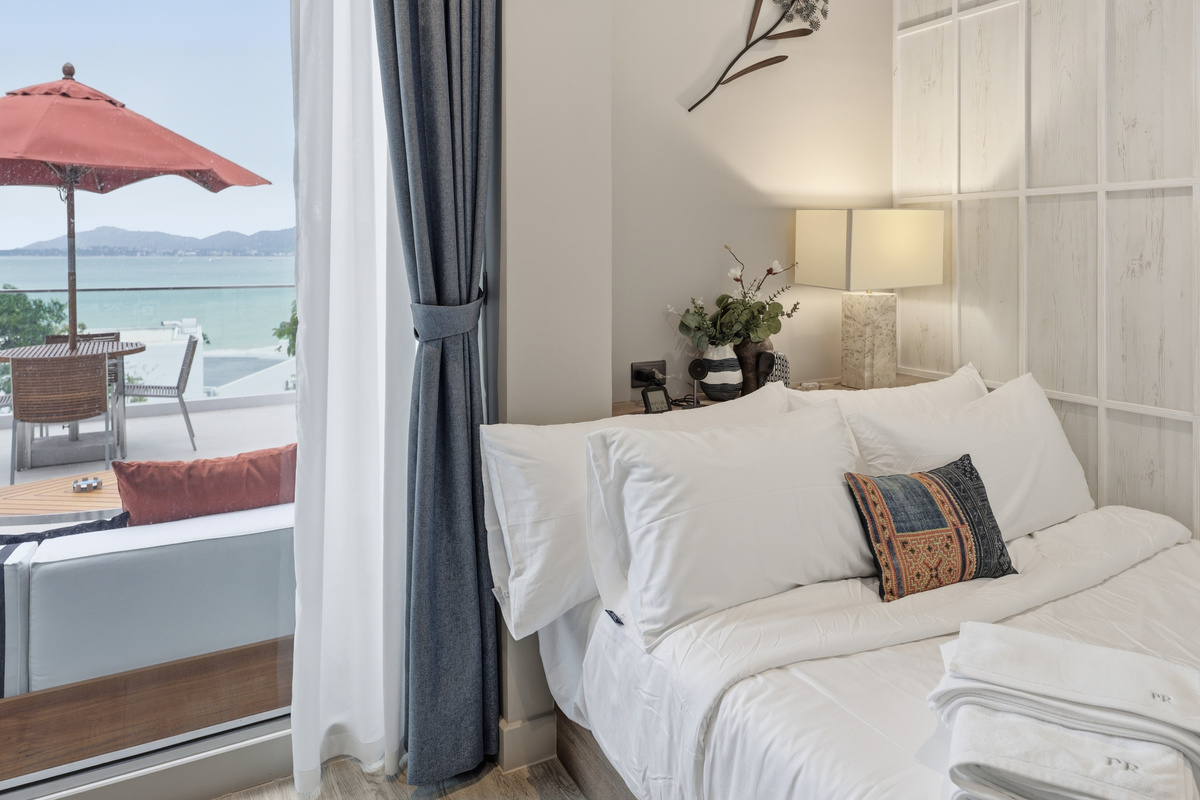 Bedroom with white linens, ocean view through glass doors, red umbrella on terrace, and coastal landscape beyond.