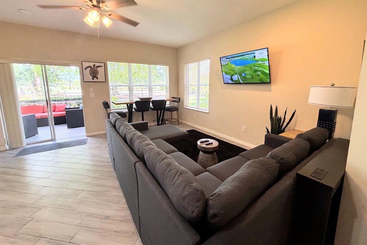 Spacious living and dining area with a large sectional, wall-mounted TV, and natural light from the patio doors.