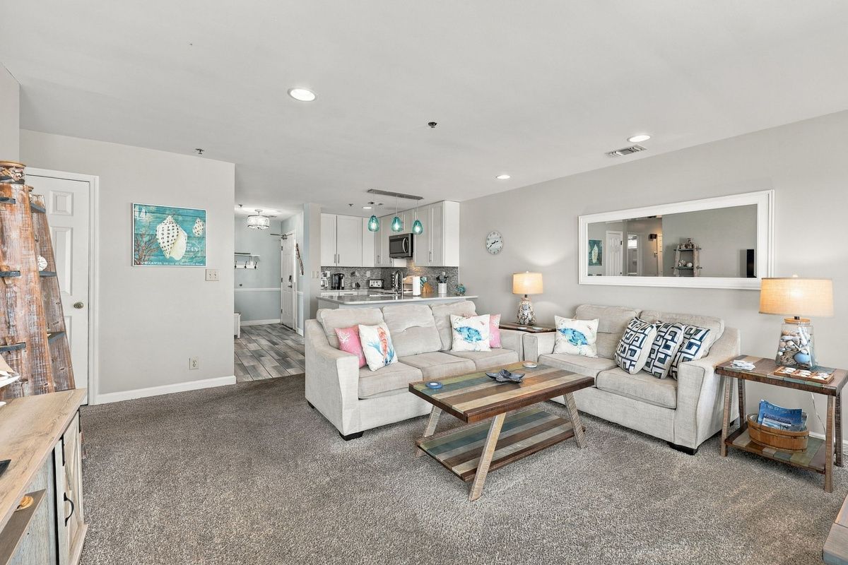 Open-concept living space with neutral sofas, a reclaimed-wood coffee table, coastal artwork, and smooth transition to the kitchen.   