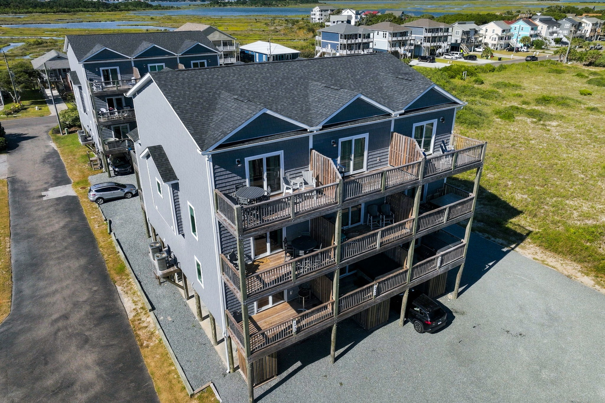 High-angle aerial perspective showing the rear exterior of the coastal townhouse complex, highlighting multi-level private decks with outdoor seating, privacy dividers, and parking beneath the elevated structure.   