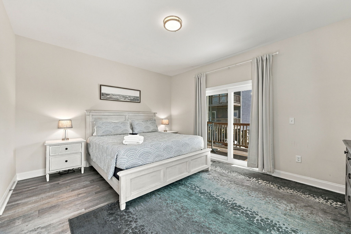 Tranquil third bedroom featuring a white wooden king bed with light blue patterned bedding, matching nightstands, a coastal area rug, and sliding glass doors leading out to a private balcony.