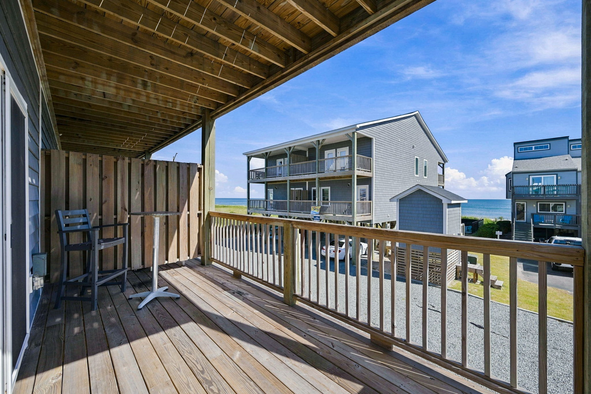 Shaded private wooden deck featuring wooden safety railings, a high-top bistro table with seating, a privacy divider wall, and views stretching out toward the ocean.   