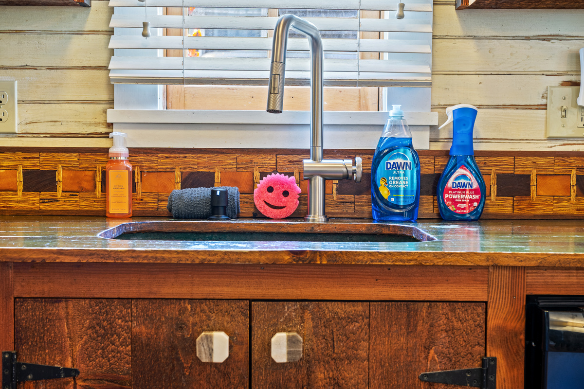 Preparing meals in the ship's galley. An upgraded high-arc faucet matches the washroom hardware, standing over a copper basin sink fully stocked with cleaning supplies and a smiling sponge beneath the window blinds.