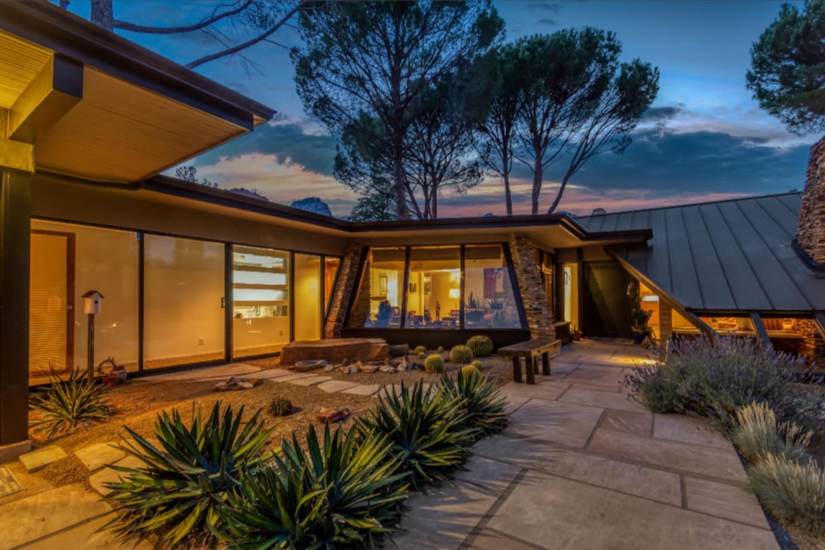 Magical evening ambiance at this mid-century retreat, where warm interior lighting glows through expansive glass walls and the desert courtyard comes alive under a stunning Sedona sunset.