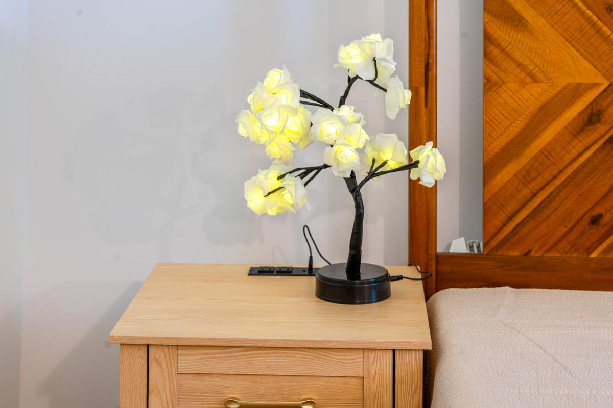 Charge your devices overnight using the handy tabletop power station with built-in ports. This detail shot highlights the light-wood nightstand in the flower room, topped with a glowing rose-branch accent lamp that provides cozy ambient light.