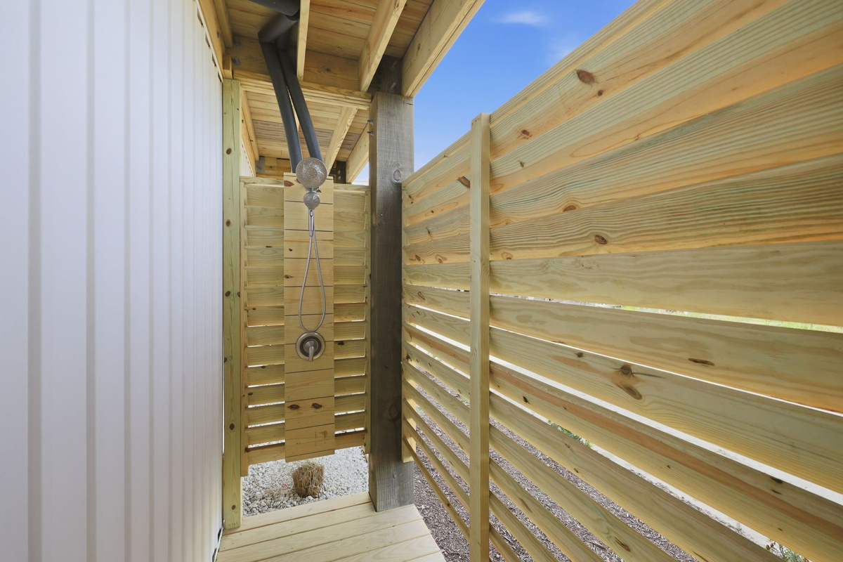Outdoor beach rinse station/shower area beneath the deck, with enclosed wooden privacy slats and mounted shower fixtures for washing off sand after beach access