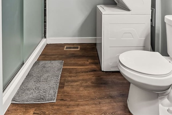 Full bathroom view showing the glass-enclosed shower, stacked washer and dryer, and durable hardwood-style flooring.