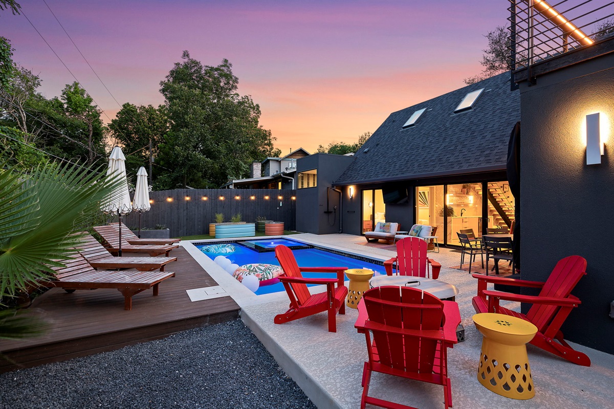 A stunning backyard oasis is illuminated at sunset, showcasing a sparkling swimming pool with a hot tub and a multi-level deck for dining and lounging.