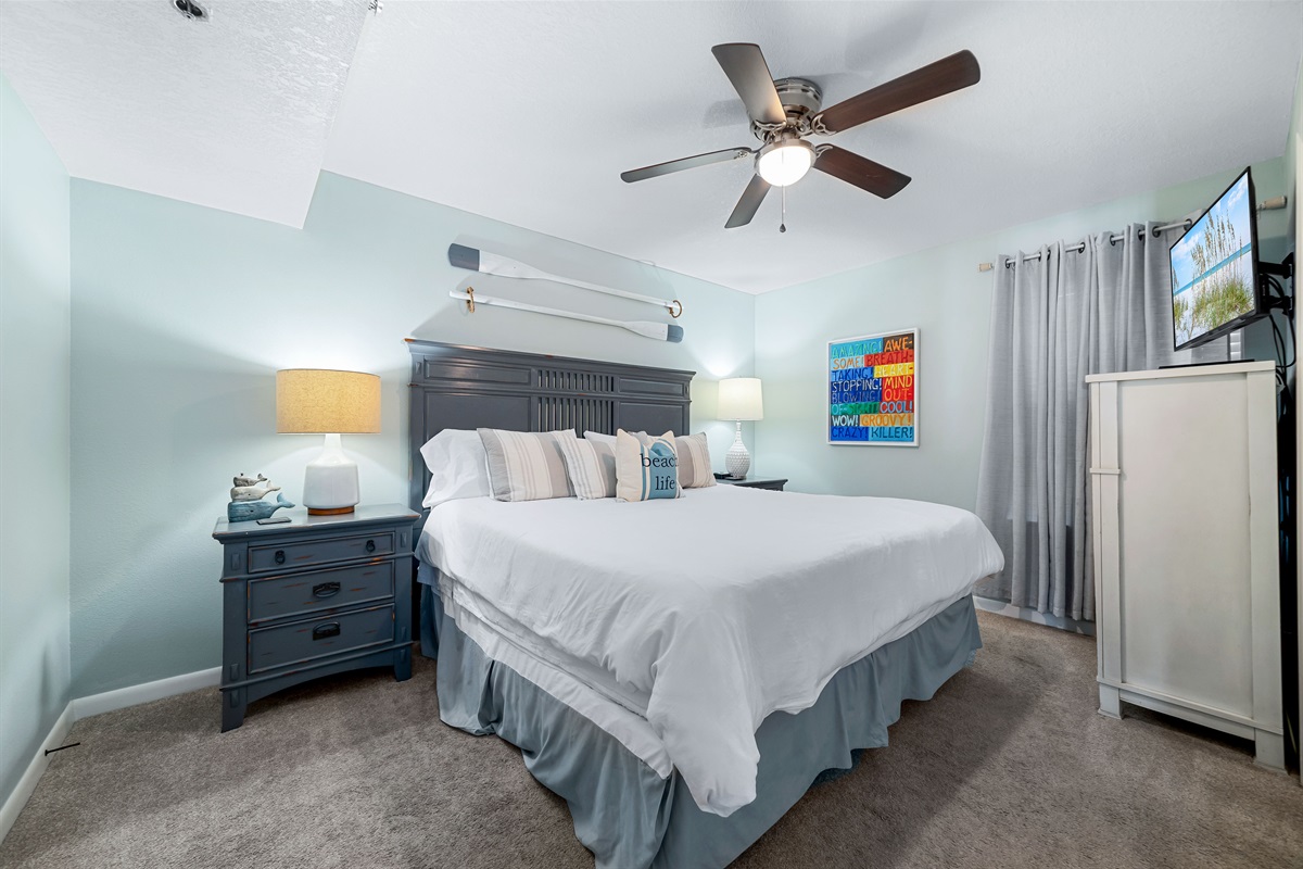 Unwind in the spacious Primary Bedroom, featuring a comfortable King-sized bed and soothing, coastal-themed decor. The room provides a peaceful sanctuary complete with a flat-screen TV and ample storage for a relaxing stay.