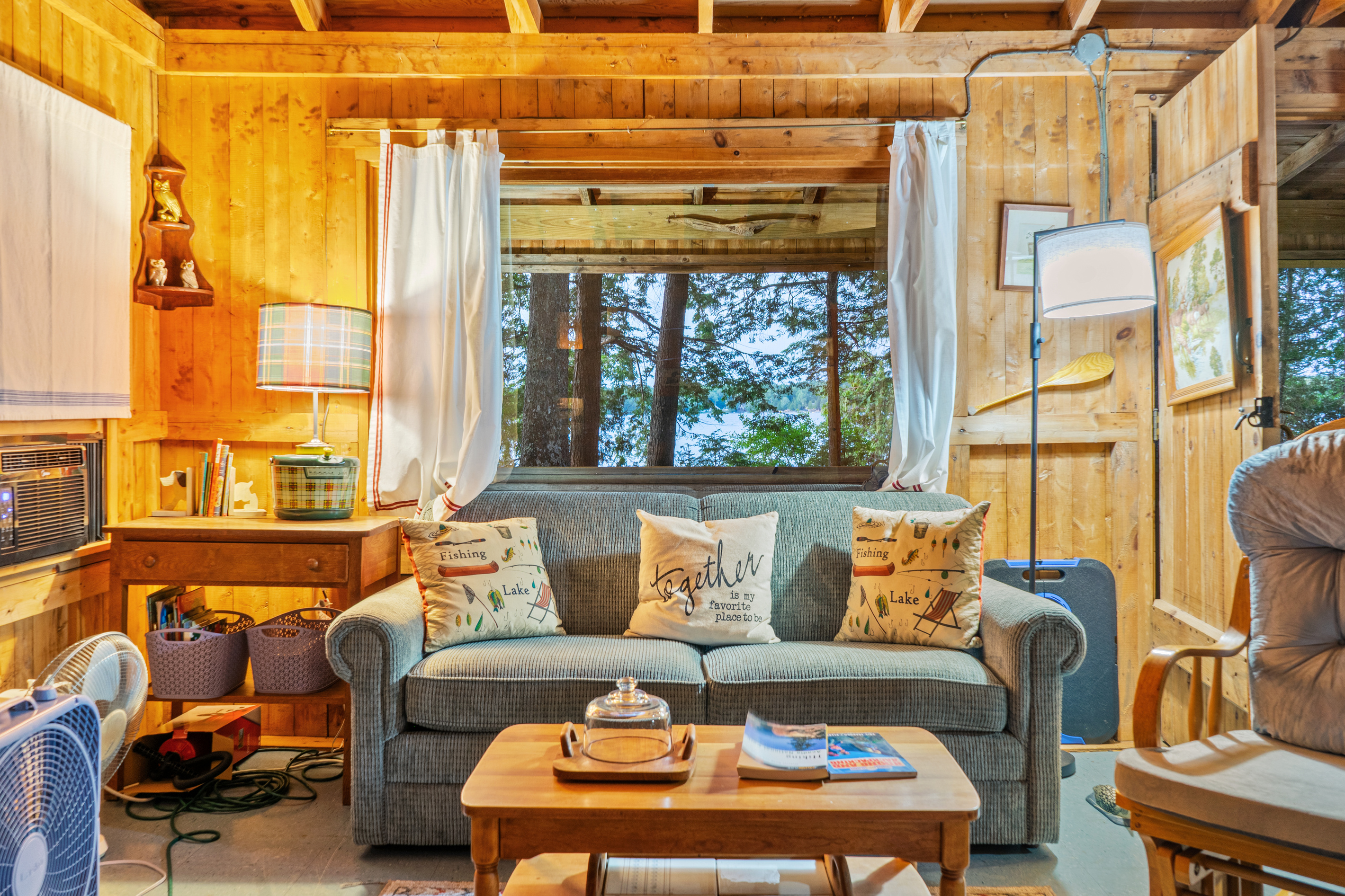 Straight-on view of the living room sofa at Loonrise Point! Big picture window frames trees and water views on Beech Hill Pond in Otis, ME. Features a comfortable sofa, window AC unit, reading table, and classic knotty pine walls for cozy evenings.