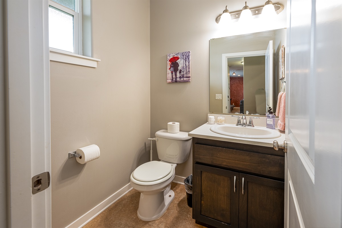 A small, well-lit bathroom featuring a white toilet, a sink with a dark wooden cabinet, a white countertop, a mirror, mounted lighting, a pink towel hanging on the wall, and a piece of wall art depicting a person with a red umbrella.
