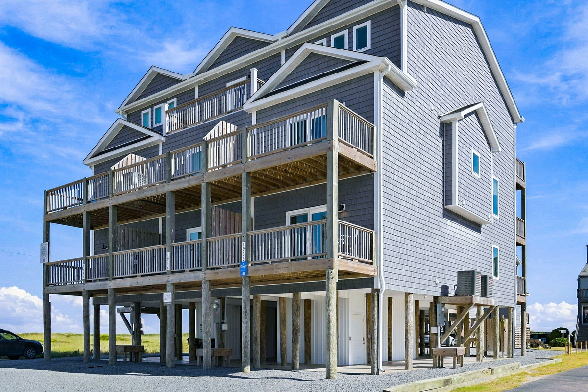 Angled exterior view showcasing the multi-story beachfront complex with coastal blue siding, extensive wrap-around wooden decks, and elevated pilings with ground-level parking under a clear blue sky.
