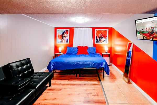 Modern Retreat: King-size Bed in Cool Blue, Accentuated by a Pop of Red Pillow, Against a Vibrant Orange and White Wall. Complete with Entertainment Center, Mirror, and Plush Theater-like Chair 🛏️🔵🔴📺
