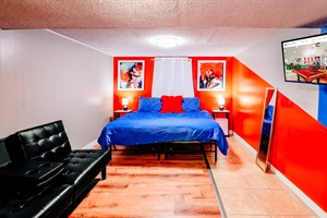 Modern Retreat: King-size Bed in Cool Blue, Accentuated by a Pop of Red Pillow, Against a Vibrant Orange and White Wall. Complete with Entertainment Center, Mirror, and Plush Theater-like Chair 🛏️🔵🔴📺