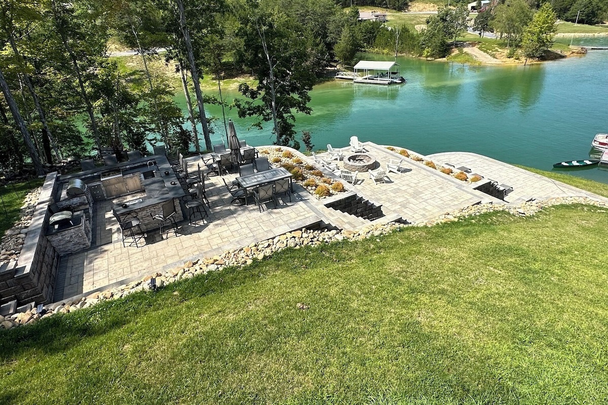 This view from the top porch looking down on the patios shows the large 16ft x 16 ft outdoor kitchen, lots of chairs to sit at the kitchen, 2 tables for eating, and an umbrella. You also see the 2nd patio with the firepit and the bottom patio for lounging.