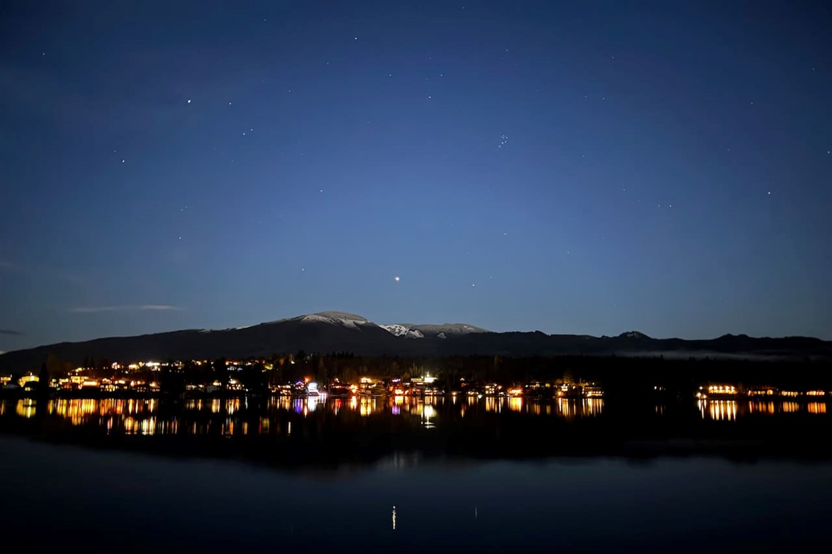 Lights from the far shoreline shimmer across the lake as the deep blue sky fades toward the horizon. The water reflects the quiet glow, creating a peaceful evening scene that feels calm, open, and still.