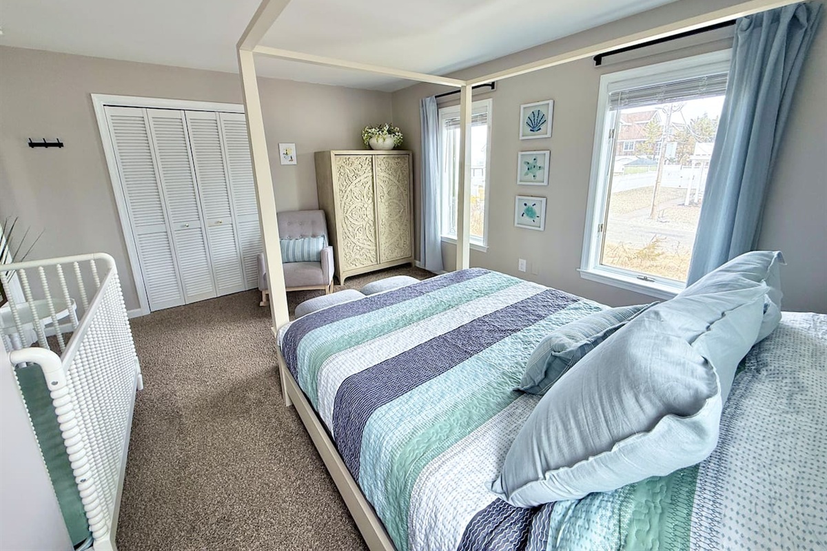 This family-friendly bedroom features a four-poster canopy bed with striped bedding and a white crib. A cozy rocking chair, decorative armoire, and coastal-themed wall art complete the serene space.