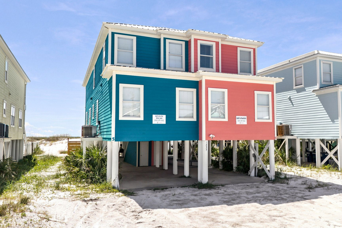 Front exterior of a vibrant beach vacation home surrounded by colorful coastal cottages and sandy pathways.