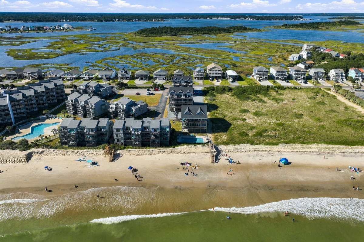 Panoramic aerial shoreline shot capturing the vibrant beachfront lifestyle, direct sandy beach access, rolling ocean waves, and the tranquil sound waters just beyond.   