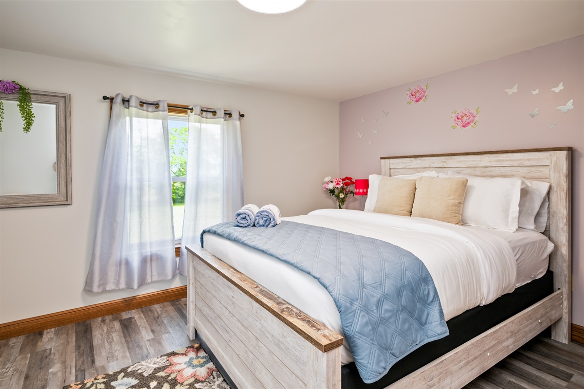 Spacious bedroom with a comfy queen bed, floral accents, and warm natural light—perfect for a restful retreat. Located on the second floor.