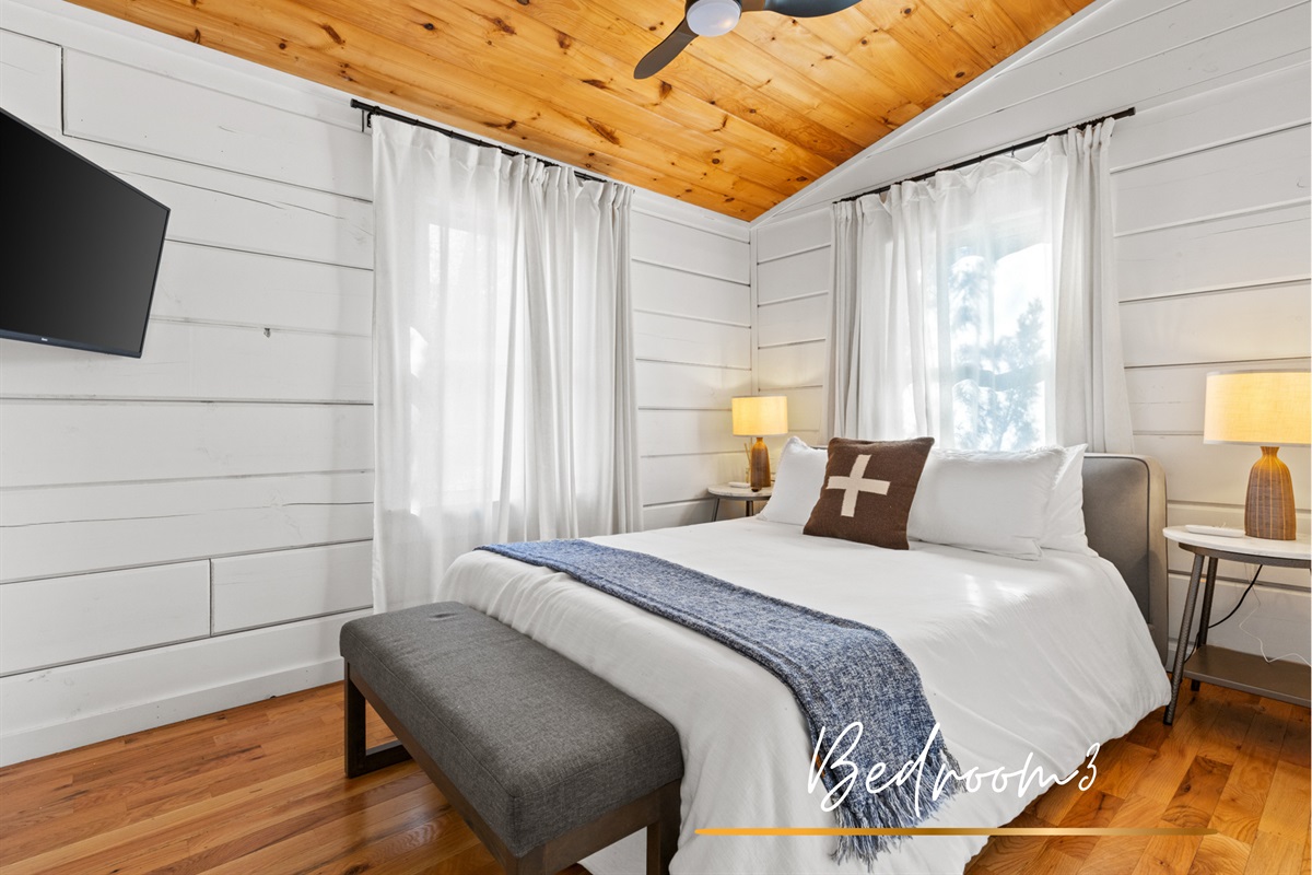 Guest bedroom offers a serene retreat with a queen-sized bed, crisp white linens, and a warm wood ceiling. Combining comfort and rustic charm.