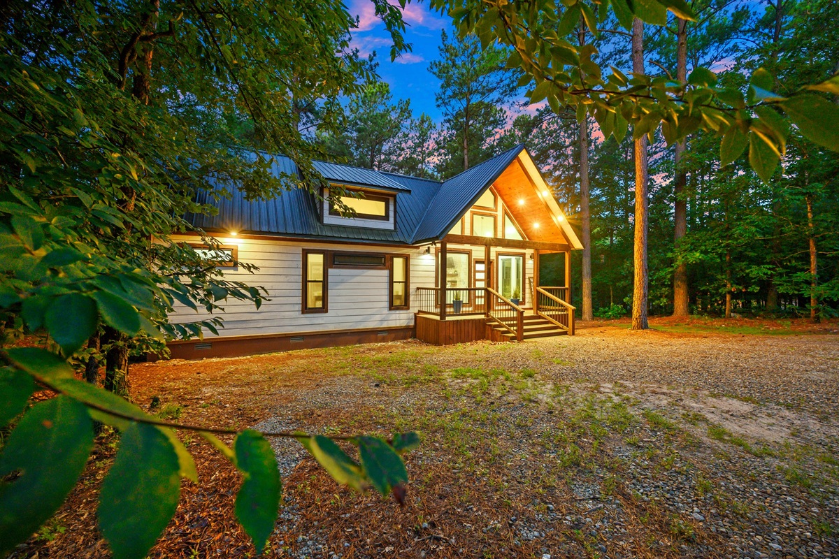 Charming Exterior: Embrace nature with warm lights and a welcoming porch amidst serene pines!