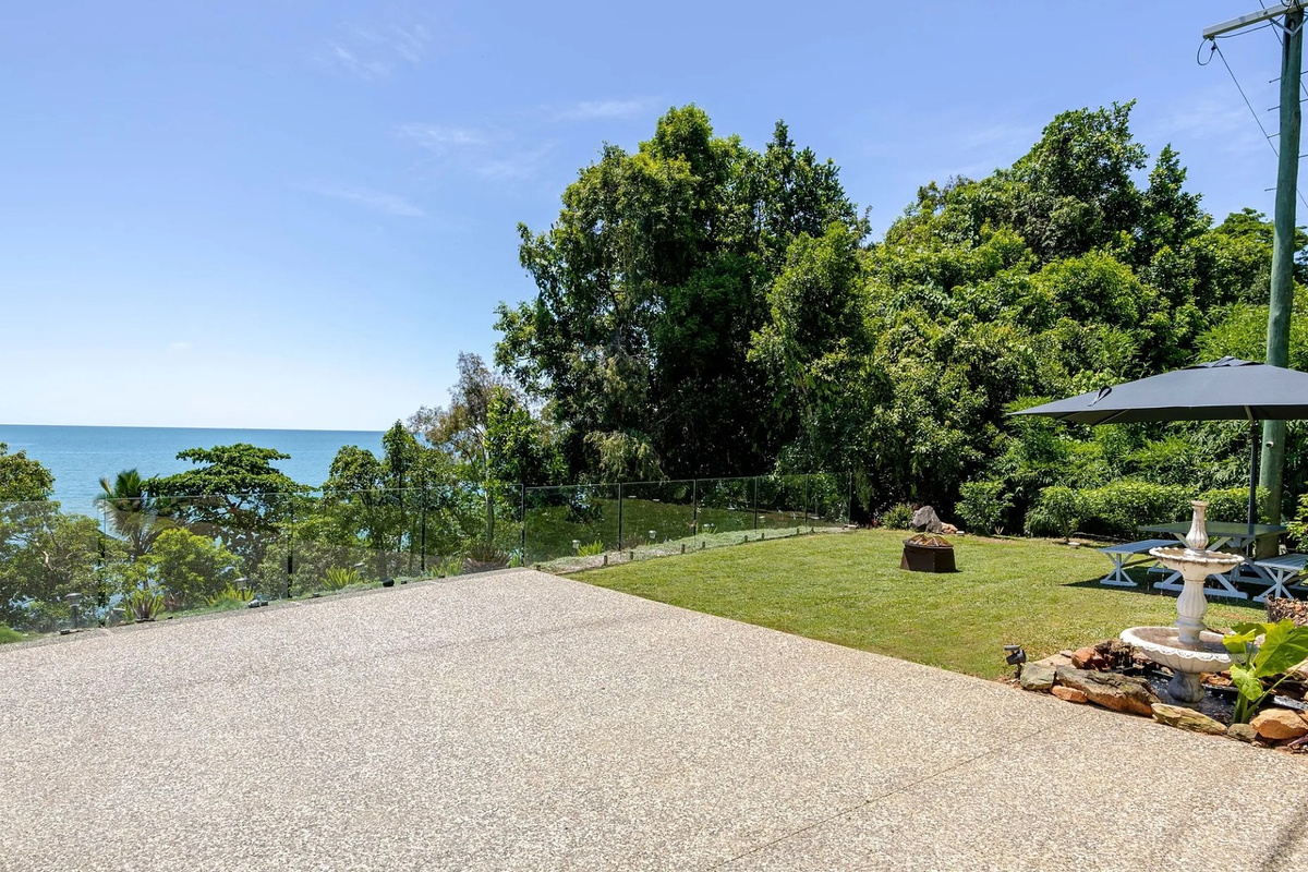 The outdoor area includes a wide driveway, lawn, glass balustrade and shaded picnic table with the ocean in view.