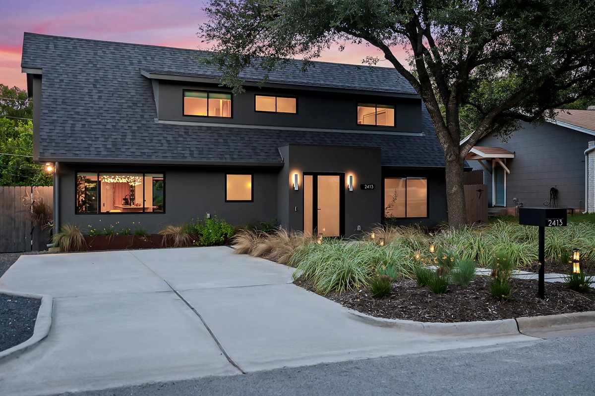 A contemporary dark gray home with a multi-level roofline, a minimalist-style front yard with a concrete driveway, and modern exterior lighting, all captured at dusk with a hint of color in the sky.