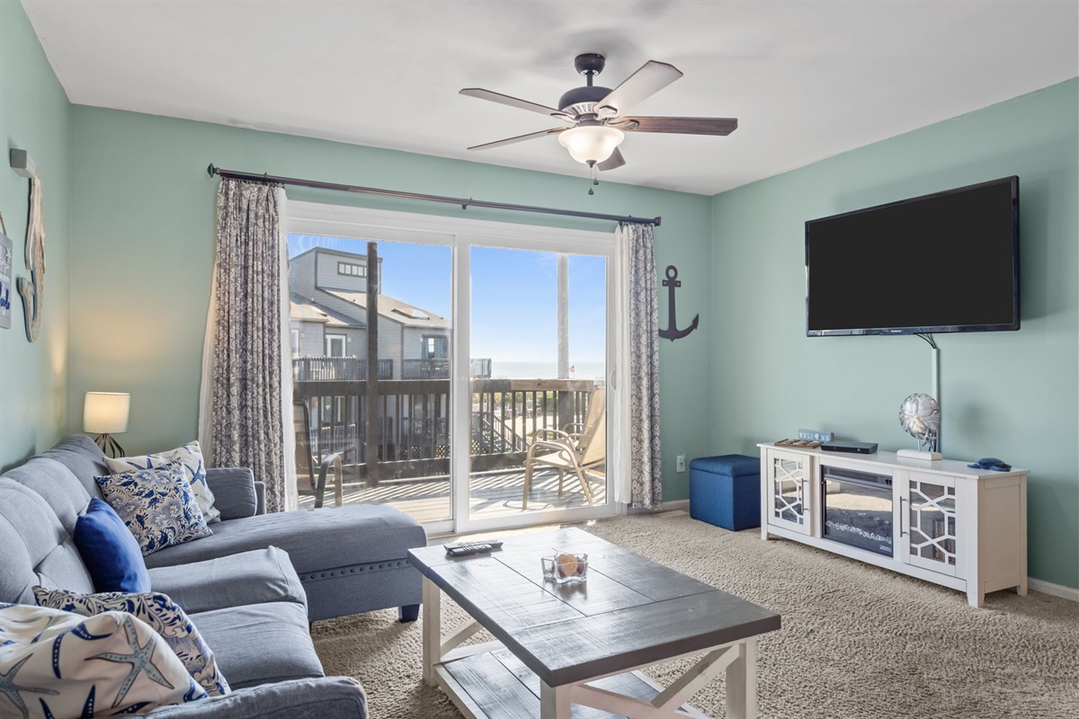 Sunlit living room with cozy seating, a large TV, and sliding doors that open to a balcony where you can catch the ocean breeze