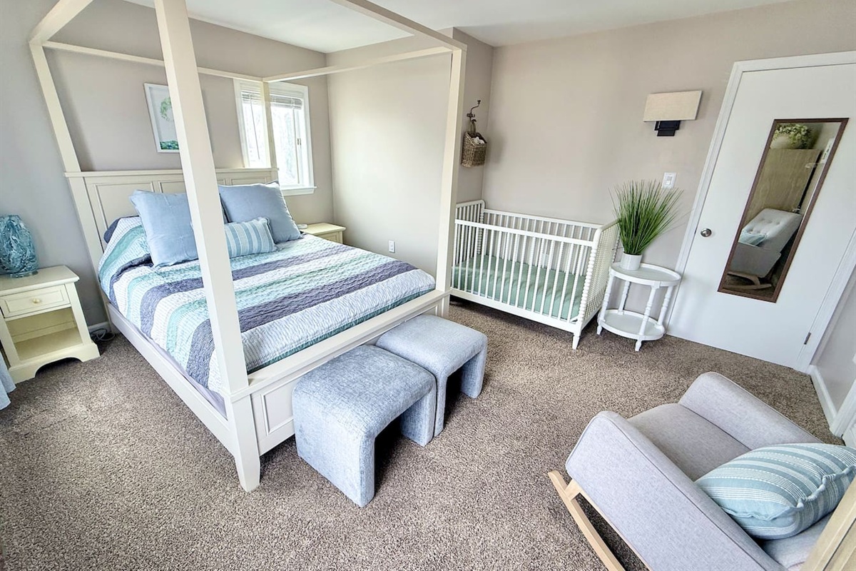 This family-oriented bedroom offers a serene retreat with a four-poster canopy bed, a white crib, and a cozy gray rocking chair. The space is completed with stylish ottomans, a side table with greenery, and a mirror.