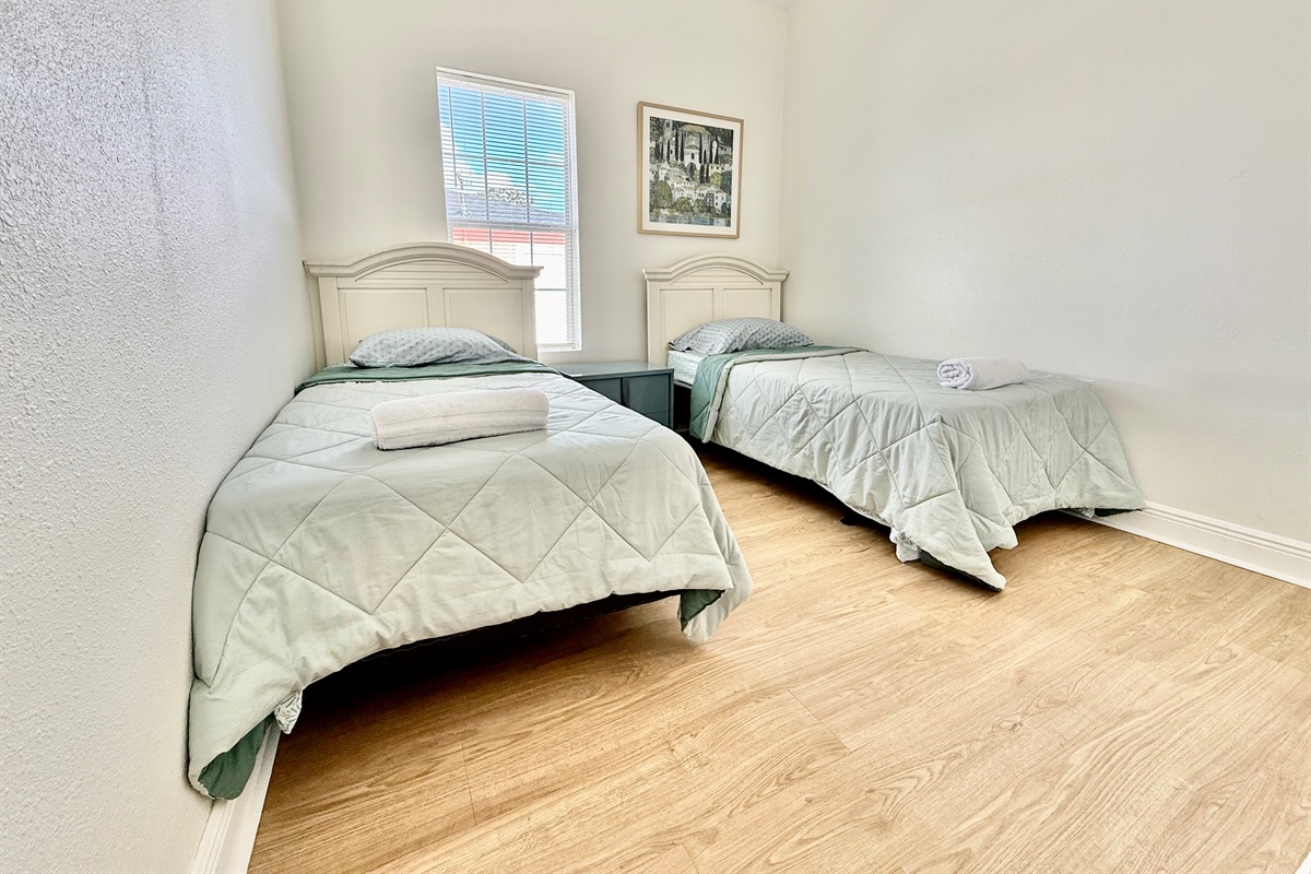 A cheerful and functional setup with twin beds, light wood flooring, and a window view to let in morning sunshine.